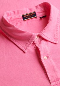 Roze poloshirt met kraag en twee knopen, met een Superdry-merklabel aan de binnenkant van de kraag.