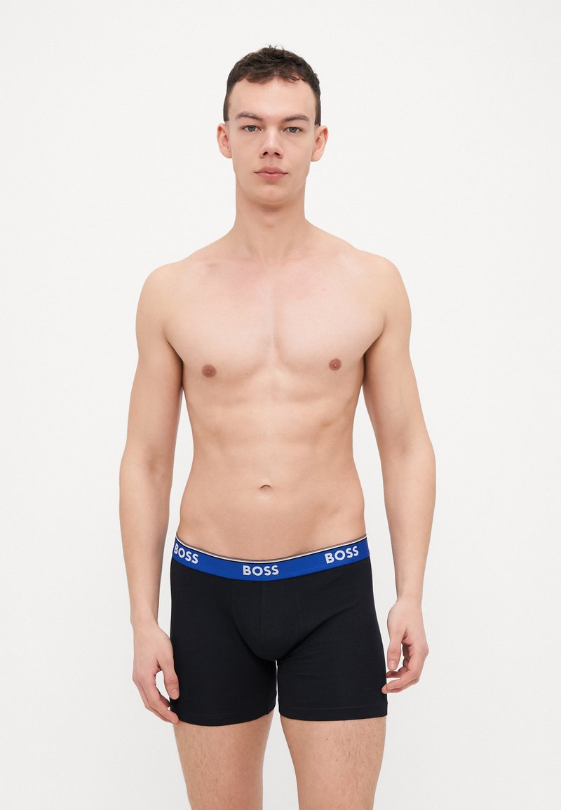Sorte boxer-short med en blå elastisk midje som har "BOSS"-logoen. Materialet ser glatt ut med en tettsittende passform. Båret av en modell.