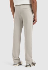 Beige broek met een ontspannen pasvorm, elastische tailleband en een klein achterzakje, gecombineerd met grijze sportschoenen met een gladde textuur.