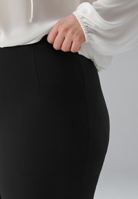 Zwarte high-waisted broek met een gladde textuur en een aansluitend ontwerp, gecombineerd met een witte, vloeiende blouse met gathering mouwen.