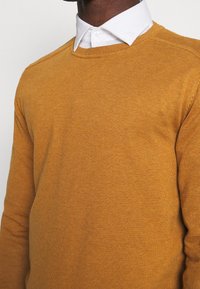 Pullover giallo senape con scollo rotondo, tessuto a coste fine e camicia colorata bianca a contrasto sotto. Tessuto a maglia liscia.
