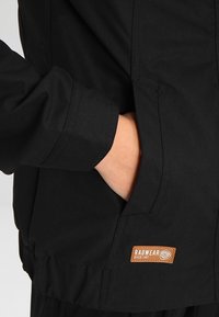 Svart jacka med strukturera yta, med en sidficka, elastiska ärmslut och en brun läderetikett som anger märket "Ragwear."