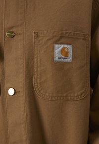 Giacca in tela marrone con una grande tasca sul petto con etichetta bianca che mostra un logo arancione e bottoni dorati lungo il davanti.