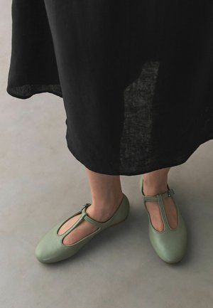 Feet wearing light green T-strap shoes paired with a black ankle-length skirt on a gray floor.