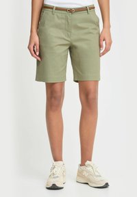 Person som har på seg olivenfargede shorts til kneet med et tynt brunt belte og beige joggesko, stående mot en ensfarget bakgrunn.