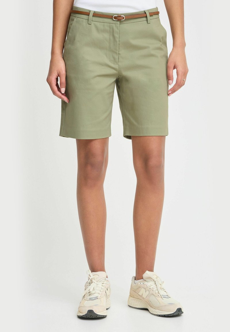 Person som har på seg olivenfargede shorts til kneet med et tynt brunt belte og beige joggesko, stående mot en ensfarget bakgrunn.