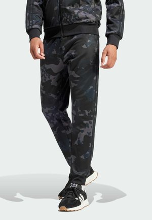 Hombre con pantalones deportivos de camuflaje en negro y gris oscuro, zapatillas negras y blancas, de pie contra un fondo claro.