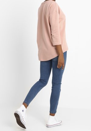 Personne portant un haut à manches longues rose clair, un jean skinny bleu et des baskets montantes blanches, debout avec une jambe légèrement levée.