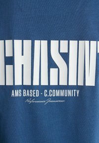 Kék pamut póló, amelynek biztos fehér szitanyomott feliratai vannak. A dizájn tartalmazza a következőket: "CHASE", "AMS BASED - C.COMMUNITY" és "Performance Jeanswear."
