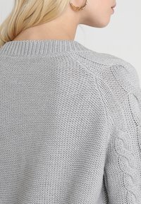 Svetle šedý pletený svetr s texturovaným vzorem. Obsahuje pružný výstřih a akcenty z kabelkového pletení na ramenou a rukávech.