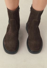 Hnědé semišové kotníkové boty s plochou černou podrážkou, bočními zipy a elegantním designem. Hladký povrch s minimálním prošitím.