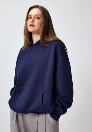 Junge Frau mit langen hellbraunen Haaren, die einen lockeren dunkelblauen Hoodie und hellbeige Hosen trägt und vor einem schlichten Hintergrund steht.