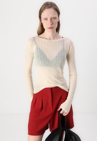 Haut à manches longues côtelé beige clair superposé sur un bralette gris, associé à un short plissé rouge ; le mannequin tient un sac noir.