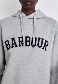Šedá mikina s kapucí vyrobená z měkké látky, s klokankou, stahovacími šňůrkami a námořnickým vyšitým logem "BARBOUR" na hrudi.