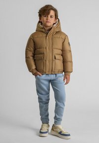 Geacă puf maro cu glugă, dotată cu două buzunare frontale și o căptușeală moale din fleece. Este combinată cu pantaloni de trening albaștri deschiși și adidași bej.