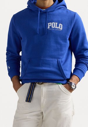 Mężczyzna ubrany w niebieską bluzę z kapturem Polo Ralph Lauren z logo na piersi, białe spodnie, paski w paski i zegarek na rękę, ręce w kieszeniach.