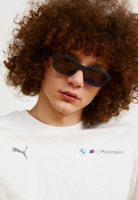 Valge puuvillane T-särk, millel on hall Puma logo ja BMW Motorsport bränding, ning seda täiendab mustad päikeseprillid, millel on ristkülikukujuline vorm ja tumendatud läätsed.