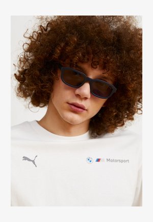 T-shirt bianca in cotone con logo grigio Puma e branding BMW Motorsport, abbinata a occhiali da sole neri con forma rettangolare e lenti oscurate.