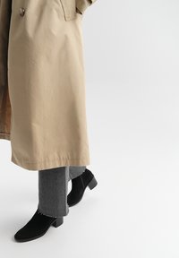Beżowy trench z gładkim wykończeniem, w połączeniu z ciemnoszarymi spodniami o strukturze oraz czarnymi zamszowymi botkami za kostkę na grubym obcasie.