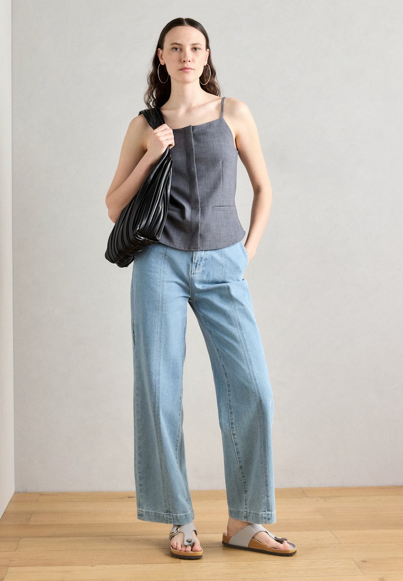 Szary bezrękawnik z cienkimi ramiączkami, niebieskie szerokie dżinsy i szare sandały. Czarna luźna torba i drewniana podłoga w tle.