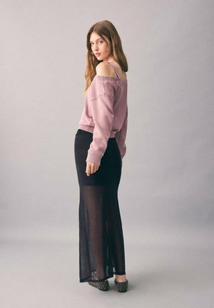 Frau mit langen Haaren, die einen pinken, schulterfreien Pullover, einen schwarzen, transparenter langen Rock und genietete schwarze Schuhe trägt, steht vor einem schlichten Hintergrund.