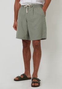 Pantalones cortos de algodón verde claro con cintura elástica y cordón. Presentan bolsillos laterales y un corte recto. Se llevan con sandalias oscuras.