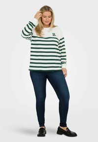 Sweat-shirt blanc à manches longues avec des rayures horizontales vert foncé, orné d'un logo sur la poitrine, associé à un jean foncé et des mocassins noirs.