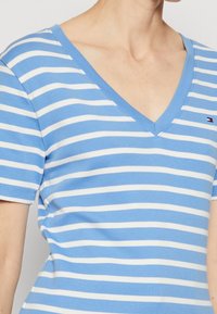 Blauwe en witte gestreepte V-hals t-shirt van katoen. Korte mouwen en een klein logo accent nabij de zoom. Zacht, standaard pasvorm.