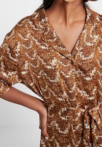 Bruin slangenprint jurk met zachte stof, korte mouwen, kraag, zijstrik op de taille en witte accenten in het patroon.