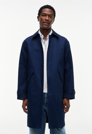 Homme portant un manteau bleu marine boutonné par-dessus une chemise blanche zippée et un jean bleu clair sur fond blanc uni.
