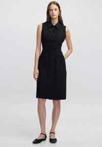Schwarzes ärmelloses Kleid mit kragenförmigem Ausschnitt, vorderem Knopfverschluss, zwei seitlichen Taschen und geradem Schnitt aus glattem Stoff.