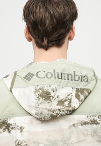 Άτομο με κοντά καστανά μαλλιά, εμφανιζόμενο από πίσω, φοράει ένα ελαφρώς πράσινο και μπεζ καμουφλάζ μπουφάν Columbia με κουκούλα.