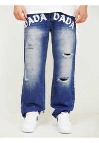 Light blue distressed jeans with prominent rips and faded detailing. "DADA" printed in white across the waistband. Worn with white sneakers.