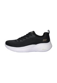 Zapatilla deportiva negra con parte superior de malla texturizada, suela blanca acolchada, diseño con cordones y un acento de logo dorado en el lateral.