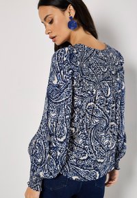 Blaue und weiße gemusterte Bluse mit weit ausgestellten Ärmeln, gerafften Details an den Schultern und einer lockeren Passform. Große blaue Ohrringe ergänzen das Design.