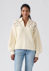 Pull en maille crème avec col en V et détail en dentelle brodée sur les épaules. Présente des manches longues bouffantes et une texture côtelée.