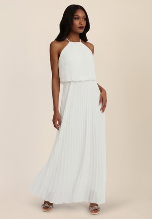 Woman wearing a white pleated halter-neck maxi dress and silver bow-adorned open-toe heels, standing against a beige background.