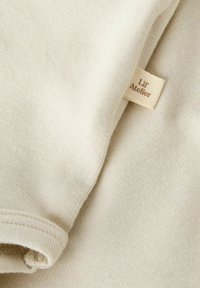 Tela de algodón color beige claro con una textura suave, que presenta una etiqueta cosida etiquetada "Lil' Atelier" en una fuente contrastante.