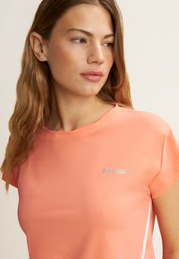 Camiseta atlética color coral hecha de tejido suave, con un cuello redondo, mangas cortas y un logo plateado en el lado izquierdo.