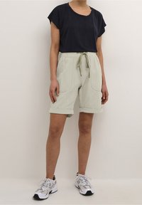 Marineblaues kurzärmeliges Oberteil, kombiniert mit hellgrünen, knielangen Shorts mit Kordelzug in der Taille und umgeschlagenen Säumen. Dazu weiße Turnschuhe.