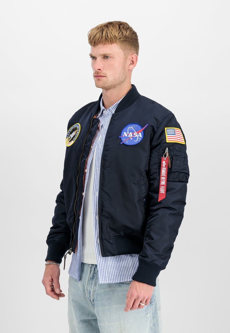 Jeune homme portant une veste bomber noire NASA avec des écussons, une chemise rayée en dessous et un jean bleu clair, tourné vers la gauche sur un fond uni.