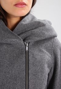 Šedá bunda z vlněné směsi s kapucí, vybavená předním zipem, texturovaným povrchem a minimalistickým designem.