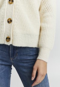 Krēmfarvet strikket cardigan med en struktureret overflade, udstyret med store, mørkebrunne knapper og ribbede manchetter, parret med blå denim jeans.