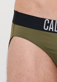 Maslinasto zeleni donji veš s crnim pojasom na kojem se nalazi bijeli tekst. Materijal izgleda glatko bez vidljivih uzoraka ili dodatnih detalja.