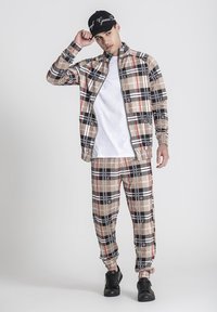 Hnedý károvaný teplákový komplet s červenými a čiernymi akcentmi; bunda na zips, nohavice s elastickým pásom, biele tričko pod ňou a čierne tenisky.