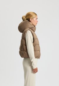 Puffiger brauner Weste mit großer Kapuze, ausgestattet mit einem steppartigen Design und seitlichen Taschen. Getragen über einem hellen, gerippten Langarm-Oberteil.