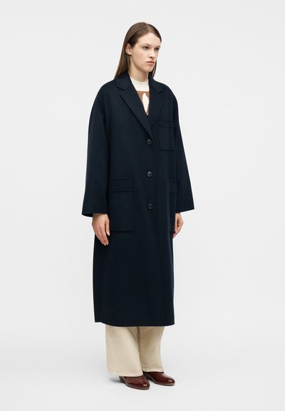Γυναίκα που φορά μακρύ, σκούρο ναυτικό παλτό με τρία κουμπιά, μεγάλες μπροστινές τσέπες, μπεζ παντελόνι και καφέ παπούτσια, στέκεται μπροστά σε λευκό φόντο.