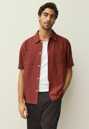 Kurzärmeliges, rostfarbenes Hemd mit zwei Brusttaschen, Knopfleiste und Kragen, aus leichtem Stoff gefertigt. Wird über einem weißen Tanktop getragen.