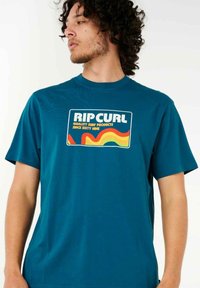 Turkis T-shirt med korte ærmer, prydet med et retro-inspireret grafisk design i orange og gult. Blødt stof med rund hals.