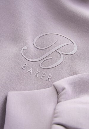 Jasnofioletowy materiał z wypukłymi literami "B" oraz "BAKER" poniżej. Tekstura jest gładka, z subtelnym połyskiem.